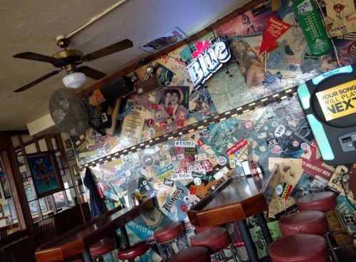A cozy bar interior featuring a colorful, sticker-covered wall and wooden tables with red stools.