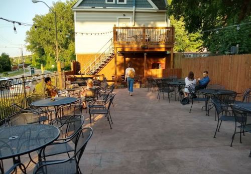 Outdoor seating area with empty tables, a woman walking, and a few people sitting at a café or restaurant.