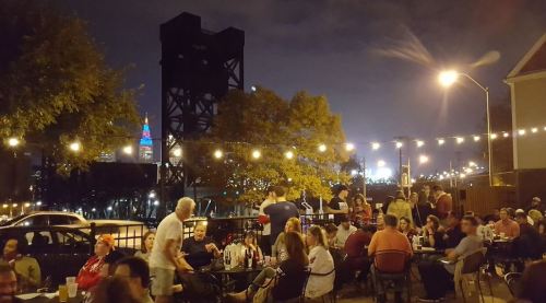 A lively outdoor gathering at night with people seated at tables, string lights overhead, and a city skyline in the background.