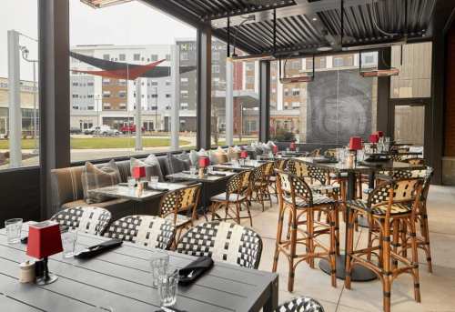 A modern restaurant patio with wicker chairs, tables set for dining, and a view of nearby buildings.