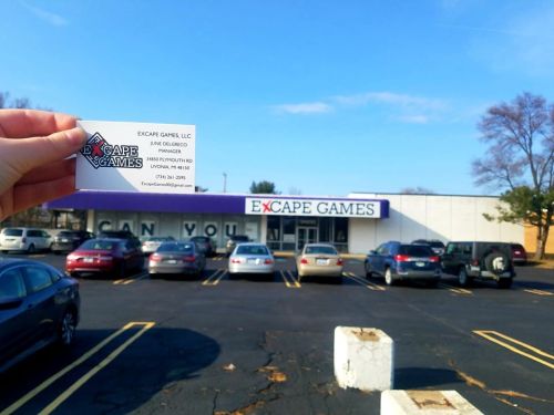A hand holds a business card in front of the Excape Games building and parking lot on a sunny day.