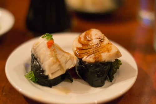 Two pieces of sushi on a white plate, featuring fish atop seaweed with garnishes and a drizzle of sauce.