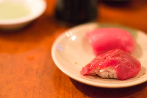 A close-up of two pieces of sushi on a small plate, featuring pink tuna atop rice, with a wooden table in the background.