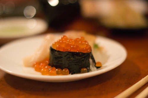A close-up of sushi on a white plate, featuring a roll topped with orange fish roe and a slice of sashimi.