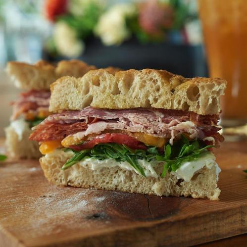 A close-up of a hearty sandwich with layers of ham, turkey, cheese, lettuce, tomato, and creamy spread on rustic bread.