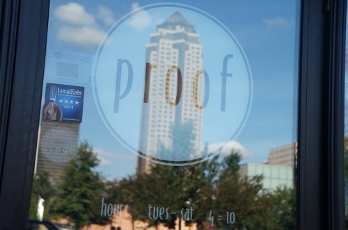 A window with the word "proof" and business hours, reflecting a city skyline in the background.