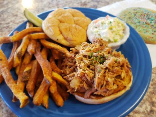 A plate with pulled pork on a bun, coleslaw, sweet potato fries, and a colorful cookie.
