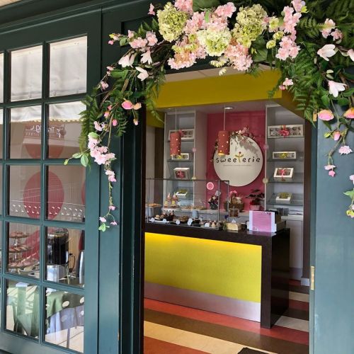 A colorful dessert shop entrance adorned with flowers and a bright sign, showcasing a variety of treats inside.