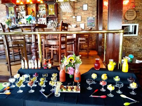 A vibrant bar setup featuring a variety of colorful cocktails, garnishes, and a floral centerpiece on a black tablecloth.