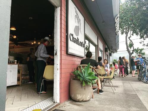 A bustling café entrance with people seated outside and a few inside, surrounded by greenery and a vibrant atmosphere.