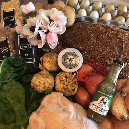 A variety of fresh groceries including lentils, potatoes, onions, greens, and a bottle of hot sauce on a wooden surface.