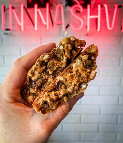 A hand holding a cookie sandwich filled with chocolate and caramel, with a neon sign in the background.