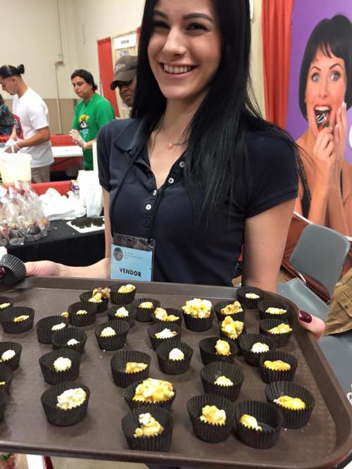 A smiling vendor holds a tray of colorful popcorn treats at a food event, with a vibrant backdrop.