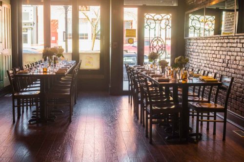 A cozy restaurant interior with wooden tables set for dining, featuring flowers and large windows letting in natural light.