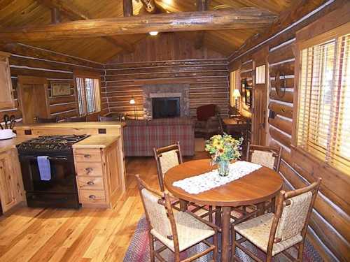 Cozy log cabin interior featuring a kitchen, dining table, and living area with a stone fireplace. Warm wooden decor.