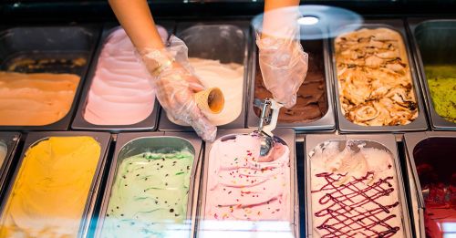 A person in gloves scoops pink ice cream from a display of various gelato flavors.