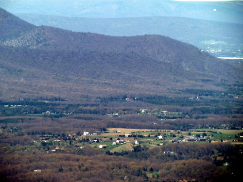 A panoramic view of rolling hills and a valley with scattered farms and a distant mountain range under a clear sky.