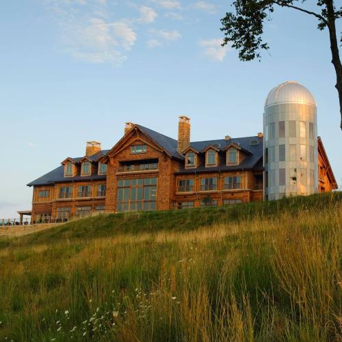 A large wooden lodge with a modern silvery silo, set against a grassy landscape and blue sky.