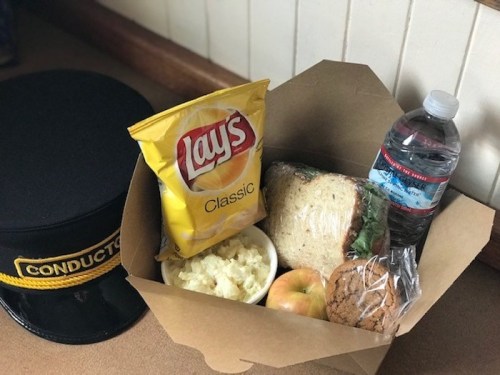 A meal box with a sandwich, chips, apple, cookie, mashed potatoes, water bottle, and a conductor's hat.