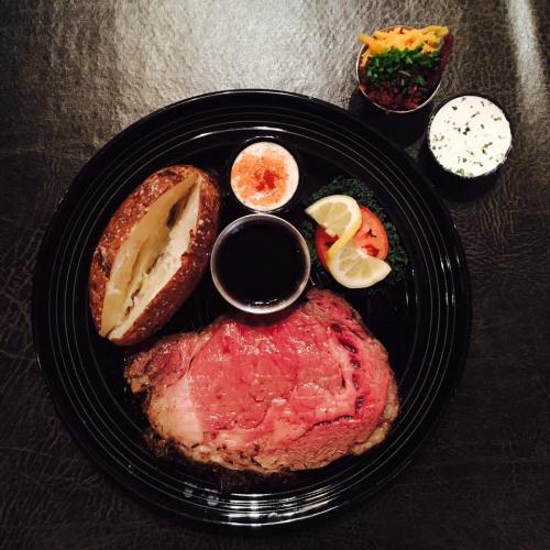 A plate featuring a juicy prime rib, baked potato, dipping sauces, and fresh vegetables with lemon slices.