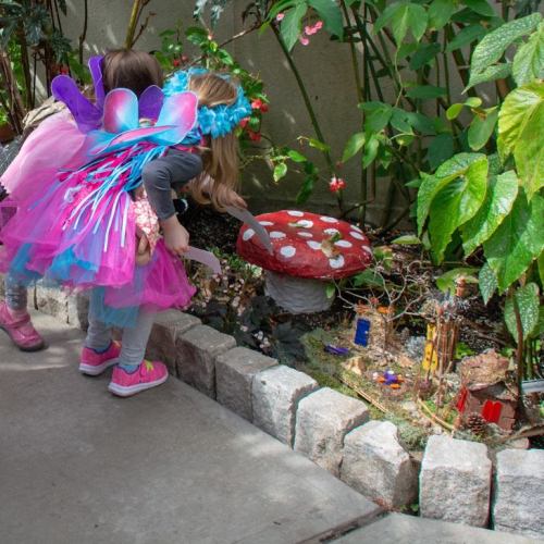 Two children in fairy costumes explore a colorful garden with a large mushroom and miniature decorations.