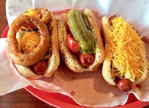 Three hot dogs in buns topped with shredded cheese, pickles, and an onion ring, served on a red tray.