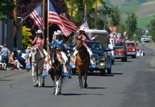 Three riders on horseback wave American flags during a parade, with spectators and vehicles in the background.