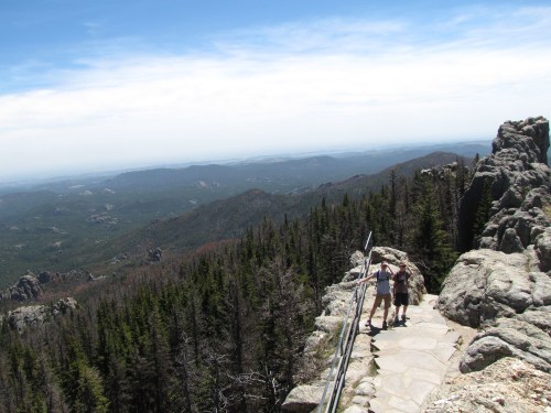 Two hikers stand on a rocky overlook, enjoying a panoramic view of mountains and forests under a clear blue sky.