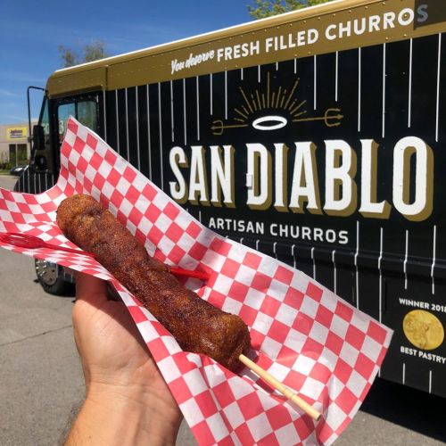 A hand holds a fresh churro on a stick in front of a food truck with "San Diablo Artisan Churros" signage.