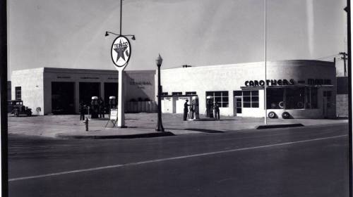 Historic gas station with vintage cars, service pumps, and people in front, featuring a Texaco sign. Black and white photo.