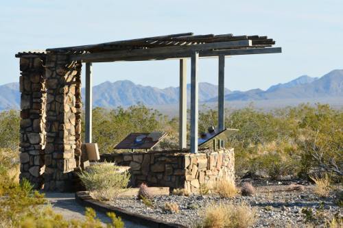 A stone shelter with a roof, surrounded by desert landscape and mountains in the background.
