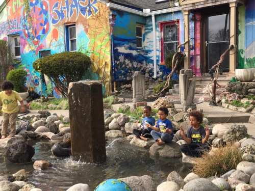 Children sit on rocks by a fountain in a colorful garden, with a vibrant mural on a nearby building.