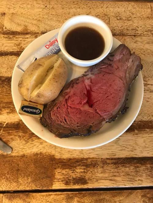 A plate with a large slice of rare prime rib, a baked potato, and a small cup of gravy.