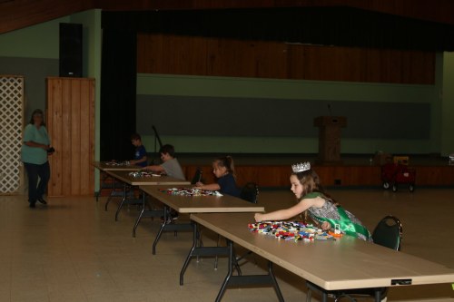 A girl in a crown sits at a table with colorful building blocks, while other children play nearby in a spacious hall.