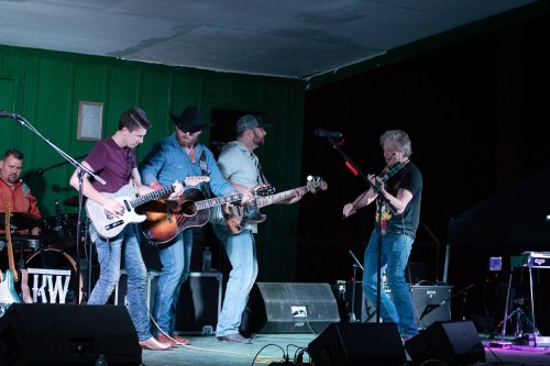 A band performs on stage, featuring guitarists and a violinist, with colorful lighting and a lively atmosphere.