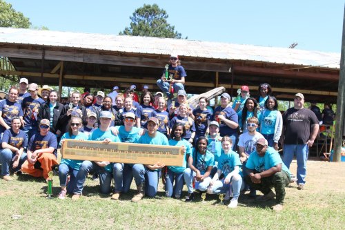 A large group of people in matching shirts poses together outdoors, holding trophies and celebrating a competition.