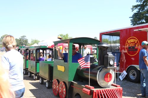 A small train ride at a fair, with children and adults enjoying the sunny day, flags waving on the train.