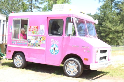 A pink ice cream truck parked in a sunny area, with a woman inside and colorful menu items displayed on the side.