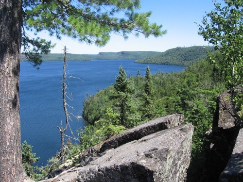 A scenic view of a calm lake surrounded by lush green forests and rocky outcrops under a clear blue sky.
