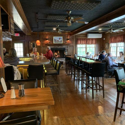 Cozy restaurant interior with wooden decor, patrons at tables, and a fireplace in the background. Warm, inviting atmosphere.