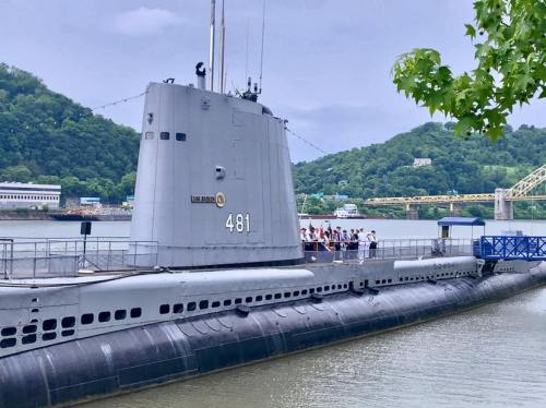 A submarine docked by a river, with people gathered on its deck and green hills in the background.