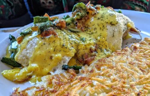 A plate of eggs topped with green vegetables and hollandaise sauce, served with crispy hash browns on the side.