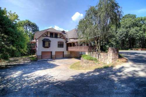 A large, two-story house with a stone foundation, surrounded by trees and a paved driveway.