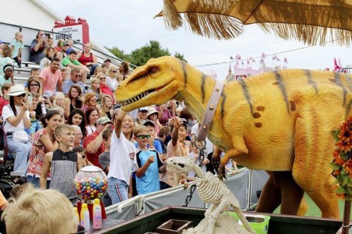 A large dinosaur puppet entertains a lively crowd of children and adults at an outdoor event.