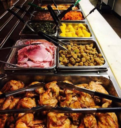 A buffet spread featuring various dishes, including meats, vegetables, and sides, with tongs for serving.
