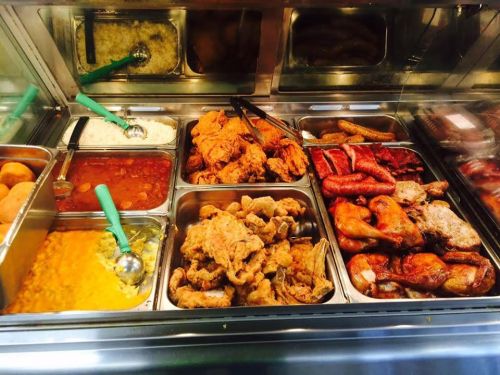 A variety of cooked dishes in metal trays, including fried chicken, sausages, rice, and beans, with serving utensils.
