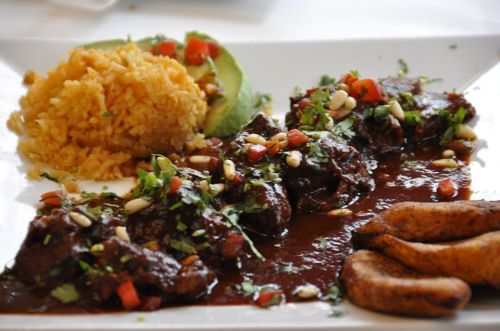 A plate of savory meat in sauce, served with rice, avocado, diced tomatoes, and fried plantains.