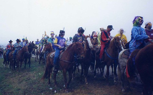 A colorful parade of people in costumes riding horses along a grassy path on a foggy day.
