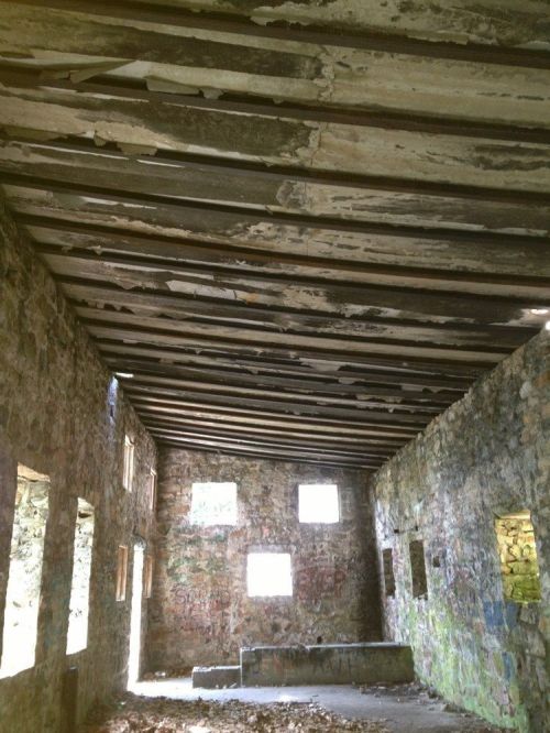 Interior of a dilapidated stone building with a wooden ceiling and several windows, showing signs of decay and graffiti.