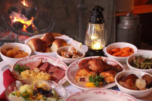 A table set with various dishes, including meats, vegetables, rolls, and a lantern, with a fireplace in the background.